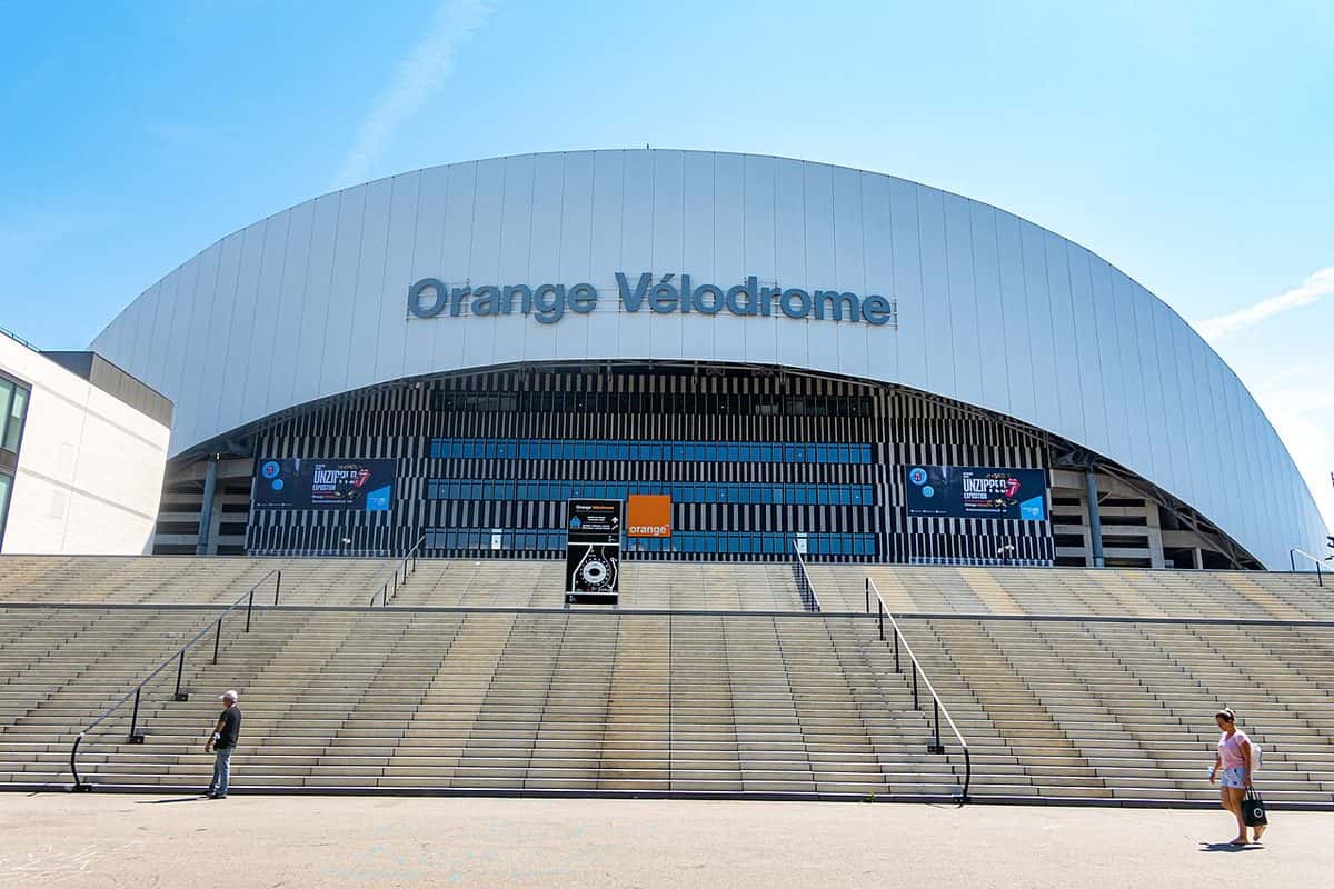 El frente del estadio Vélodrome de Marsella, con su techo blanco ondulado y la escalinata de la entrada bajo un cielo azul; foto de archivo de julio de 2021 (foto: Bernard Ddd, Wikimedia Commons, licencia CC BY-SA 2.0)