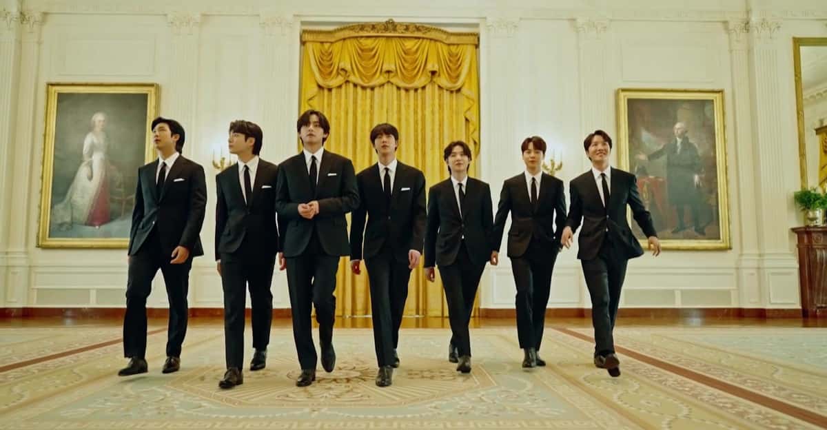 Los siete integrantes de BTS, de traje negro, caminan por un salón de la Casa Blanca con cortinas doradas y cuadros de época en mayo de 2022 (foto de archivo)